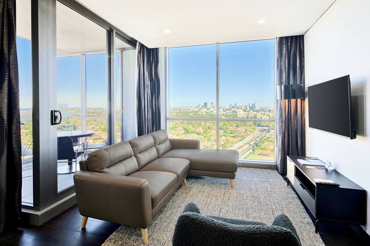 A modern living room with a sectional sofa, a flat-screen TV, and a city view through large windows.