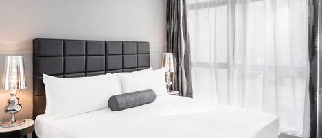 A modern hotel room with a large bed, a nightstand with a lamp, and sheer curtains allowing natural light.