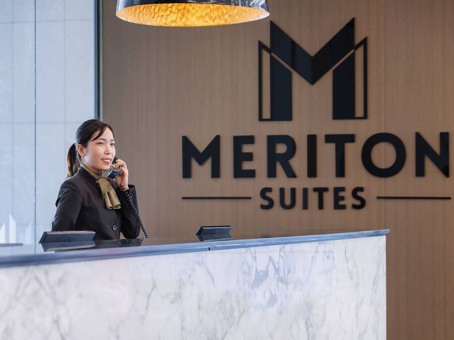 Una receptionist alla reception di Meriton Suites, con un bancone in marmo e un grande cartello con la scritta 'Meriton Suites' sullo sfondo.