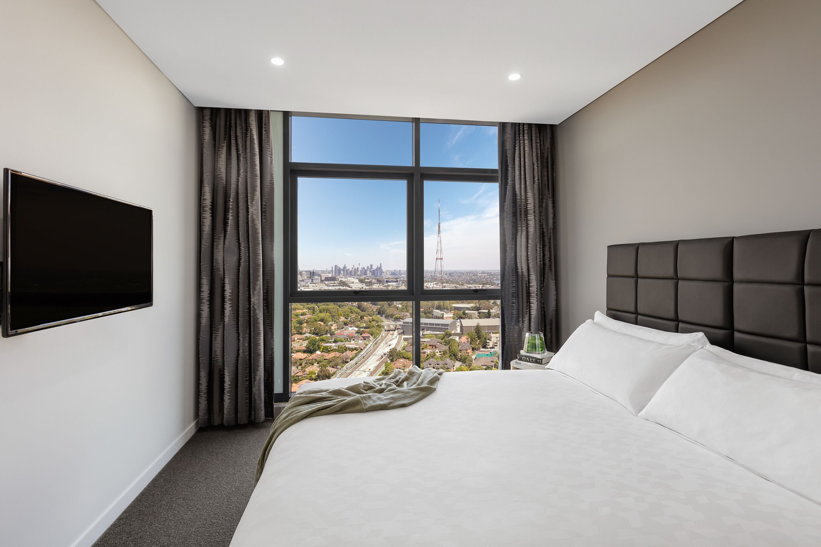 A modern hotel room with a large bed, a flat-screen TV, and a city view through large windows.