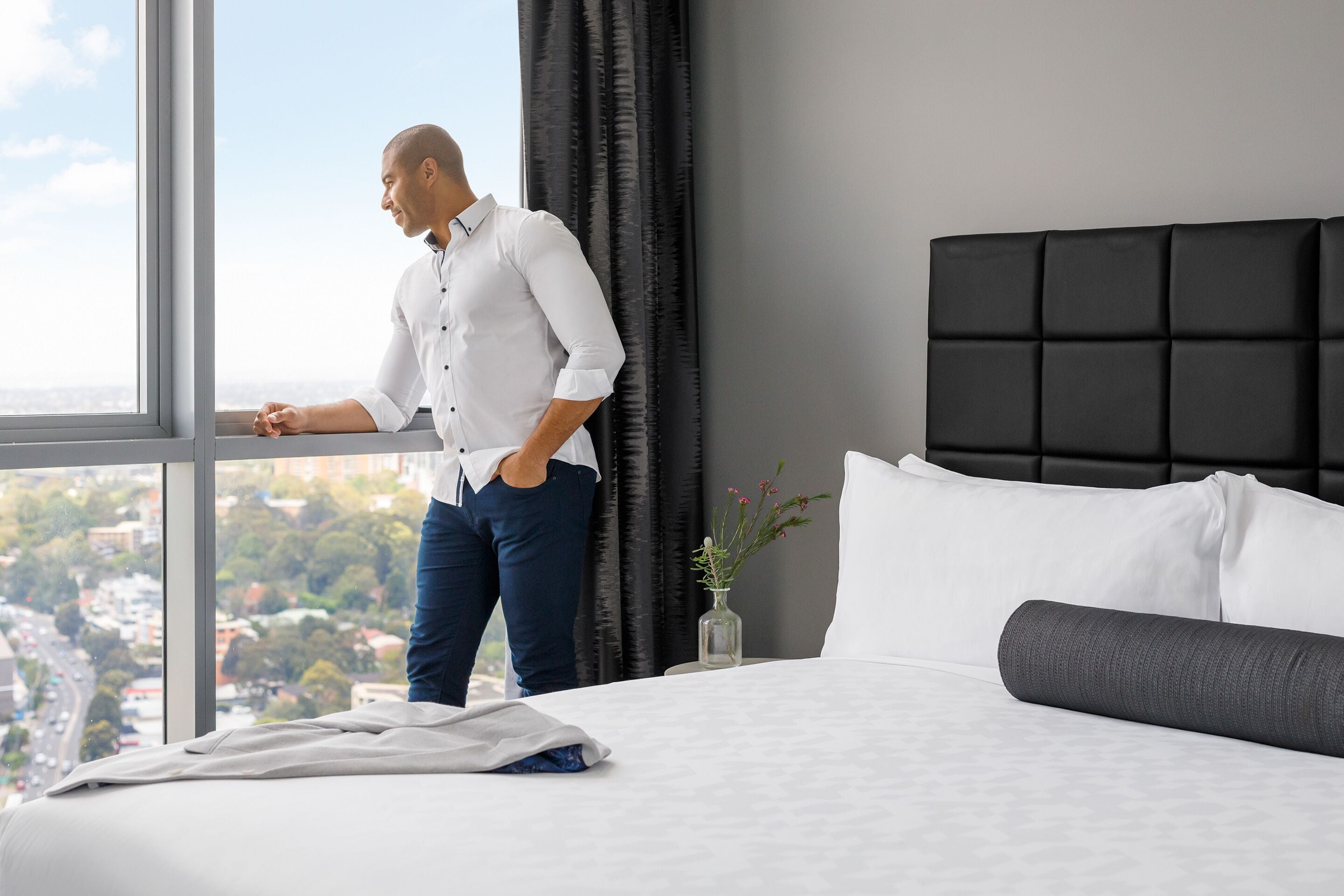 A hotel room with a large window, a bed with white bedding, a black headboard, and a small vase with flowers on the nightstand.
