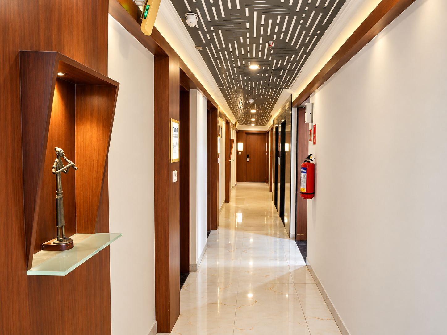 A hotel corridor with wooden paneling, a glass shelf with a brass statue, a fire extinguisher, and a ceiling with linear light fixtures.