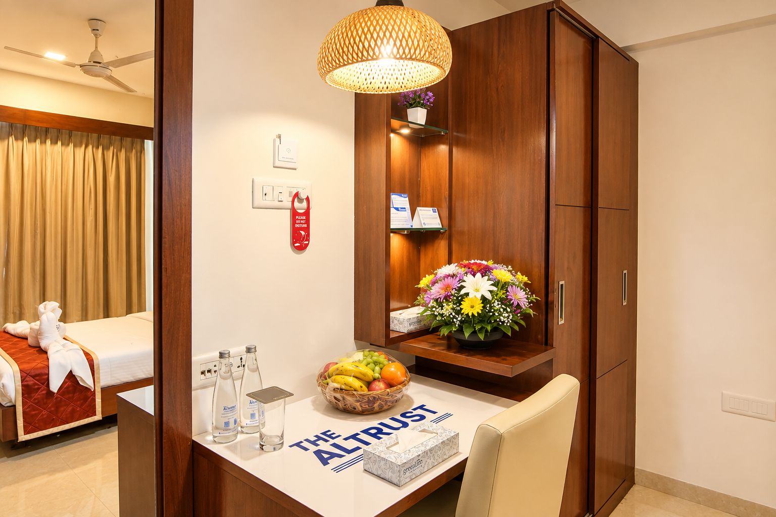 Una habitación de hotel con una cama, un escritorio con frutero, un florero, una botella de agua y un cartel que dice 'The Altrust'.