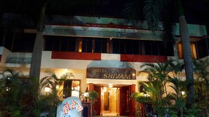 Hotel anomenat 'HOTEL SHIVAM' amb una placa, entrada i palmeres als voltants.