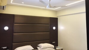 A hotel room with a modern design, featuring a large padded headboard, a ceiling fan, recessed lighting, and a bedside table with a lamp. - Hotel Shivkrupa (Pune)