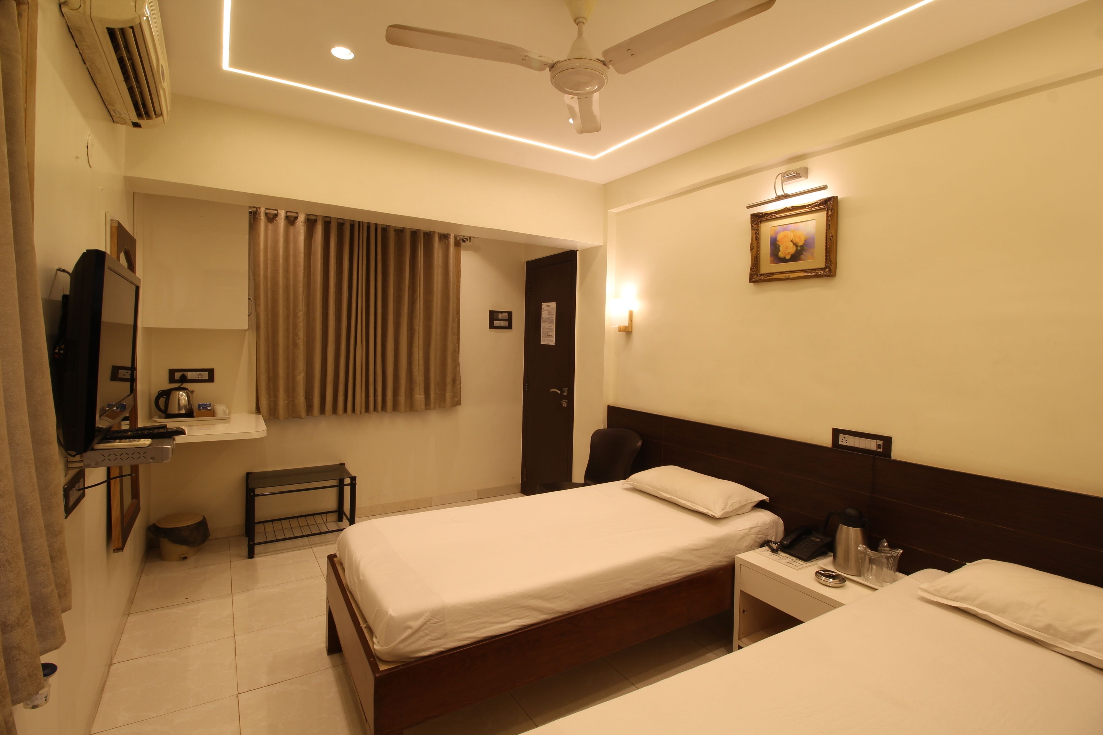 A hotel room with two beds, a desk, a chair, an air conditioning unit, a ceiling fan, a picture on the wall, and a window with curtains.