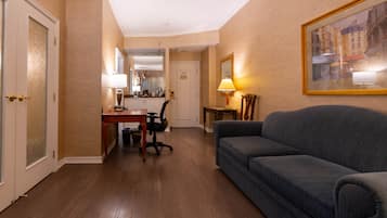 Une chambre d’hôtel avec un plancher en bois, un canapé gris, un bureau avec une chaise, une lampe et un tableau encadré au mur.