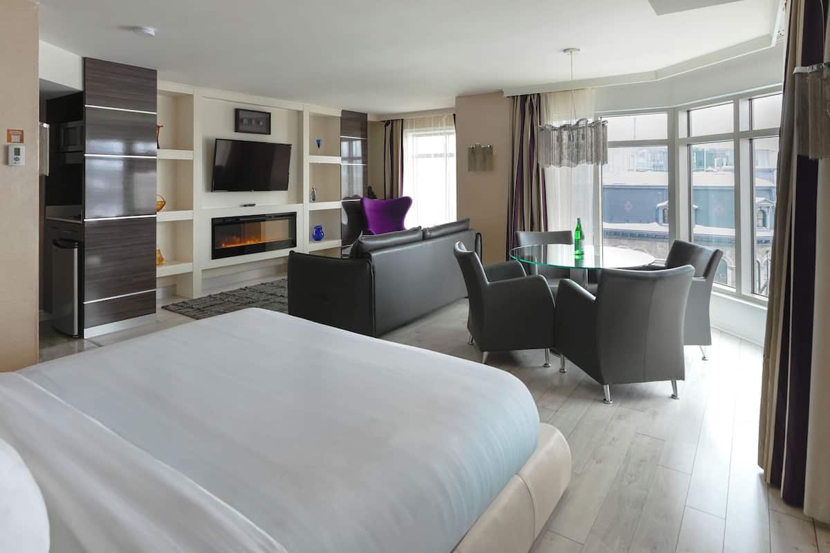 Ett hotellrum med en stor säng, en sittgrupp med ett glasbord, en platt-TV, en öppen spis och utsikt över staden genom stora fönster.