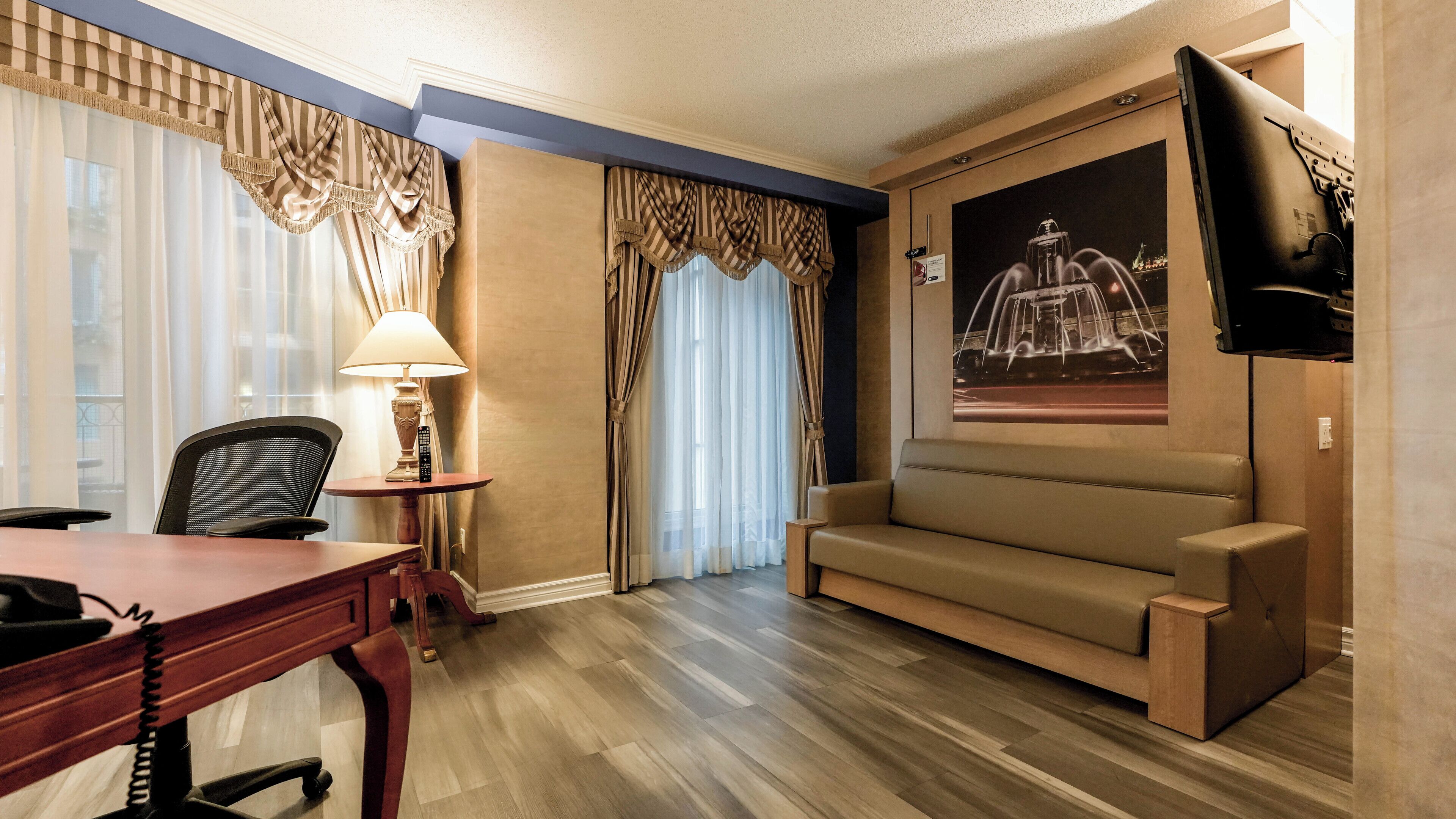 Habitación de hotel con escritorio de madera, una silla, un sofá, televisor montado en la pared y una ventana con cortinas.