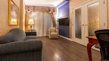 Un salon comprenant un canapé, un fauteuil et un téléviseur fixé au mur. On y trouve une porte avec des panneaux en verre dépoli et un rideau agrémenté d’une lambrequin à motifs.