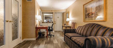 A hotel room with a wooden floor, a striped sofa, a desk with a chair, and a dining area.
