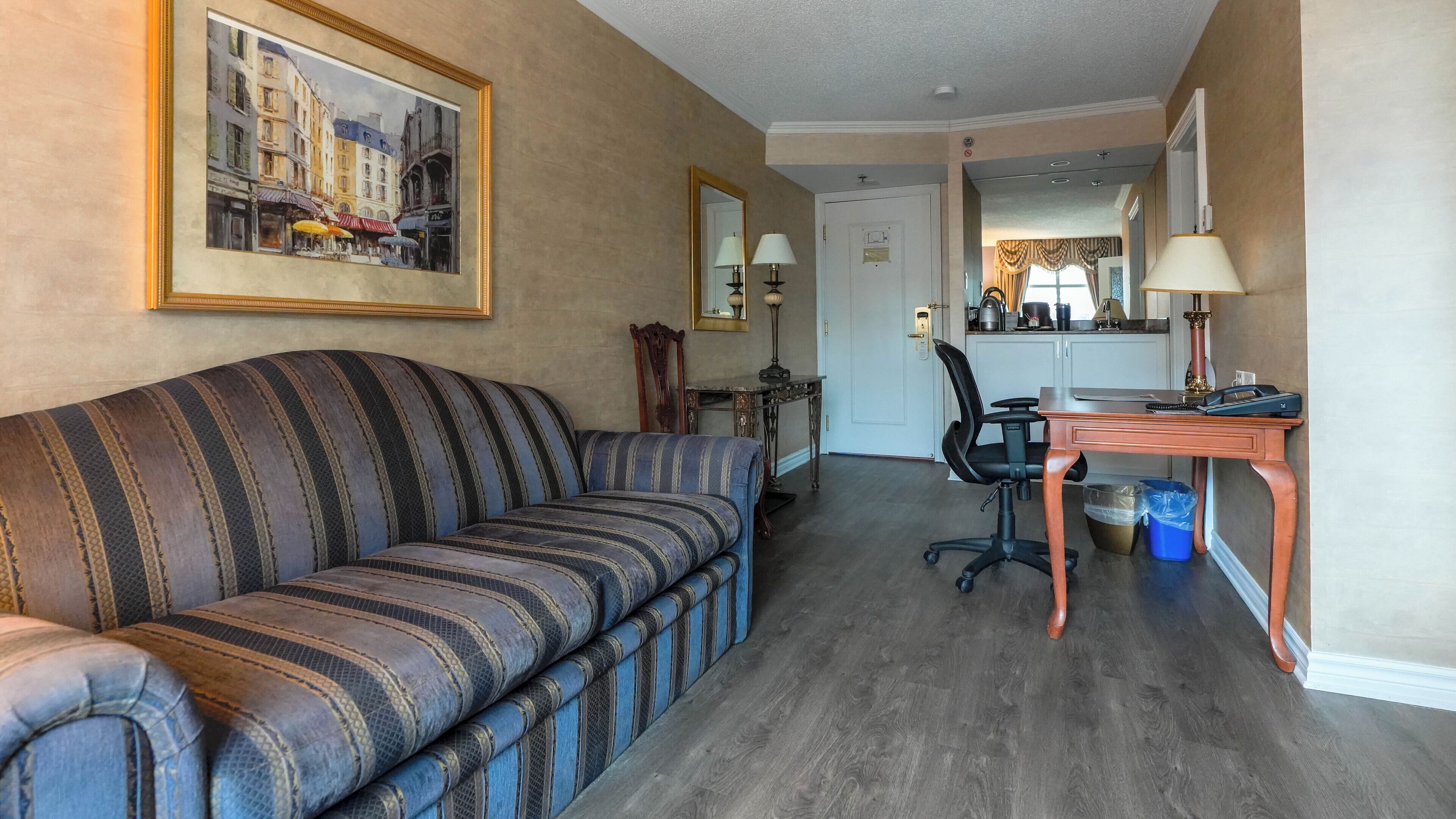 A hotel room with a striped sofa, a desk with a chair, a mirror, and a framed painting on the wall.