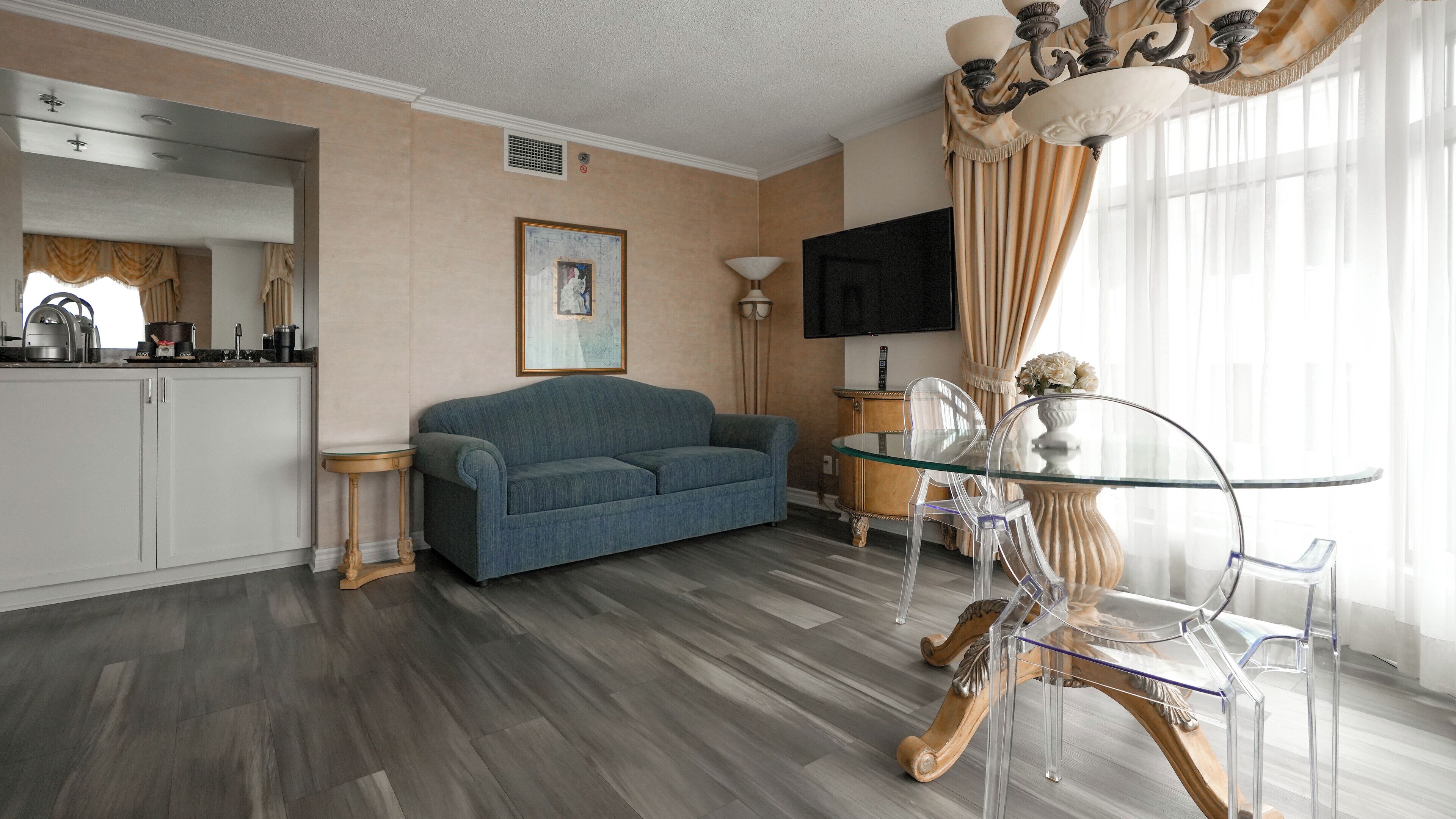 Ein modernes Wohnzimmer mit Sofa, einem Glastisch mit Stühlen, einer Küchenzeile und einem großen Fenster mit Vorhängen.