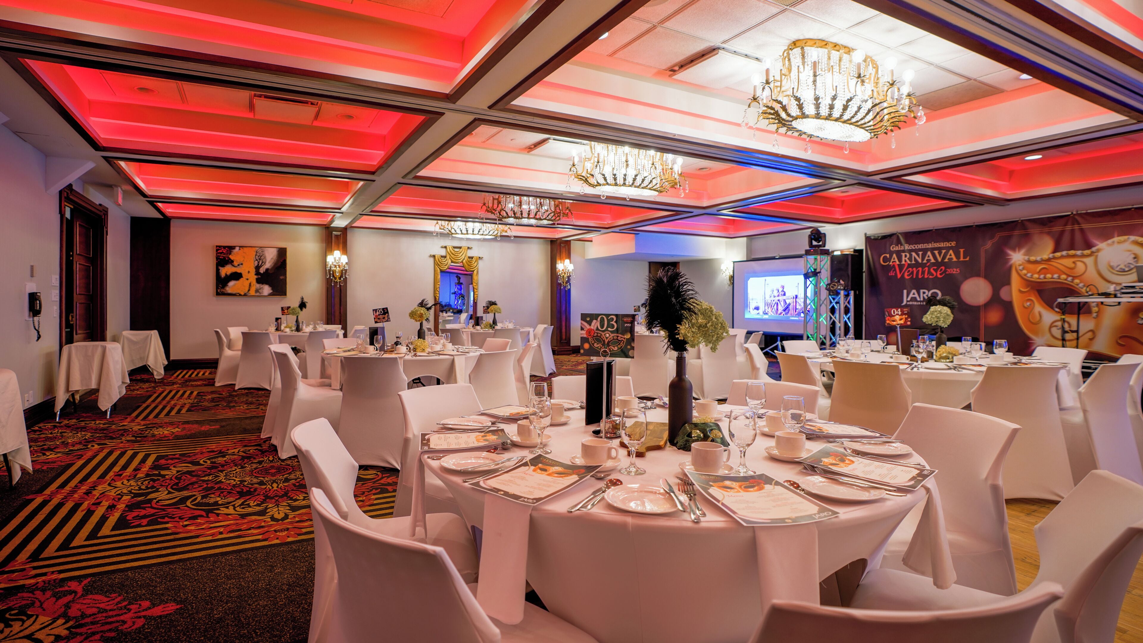 A banquet hall with round tables, white chairs, and chandeliers. Tables are set with plates, glasses, and cutlery. A large screen displays an event.