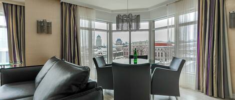 Quarto de hotel moderno com área de refeições no canto, grandes janelas com cortinas e vista para a paisagem urbana.