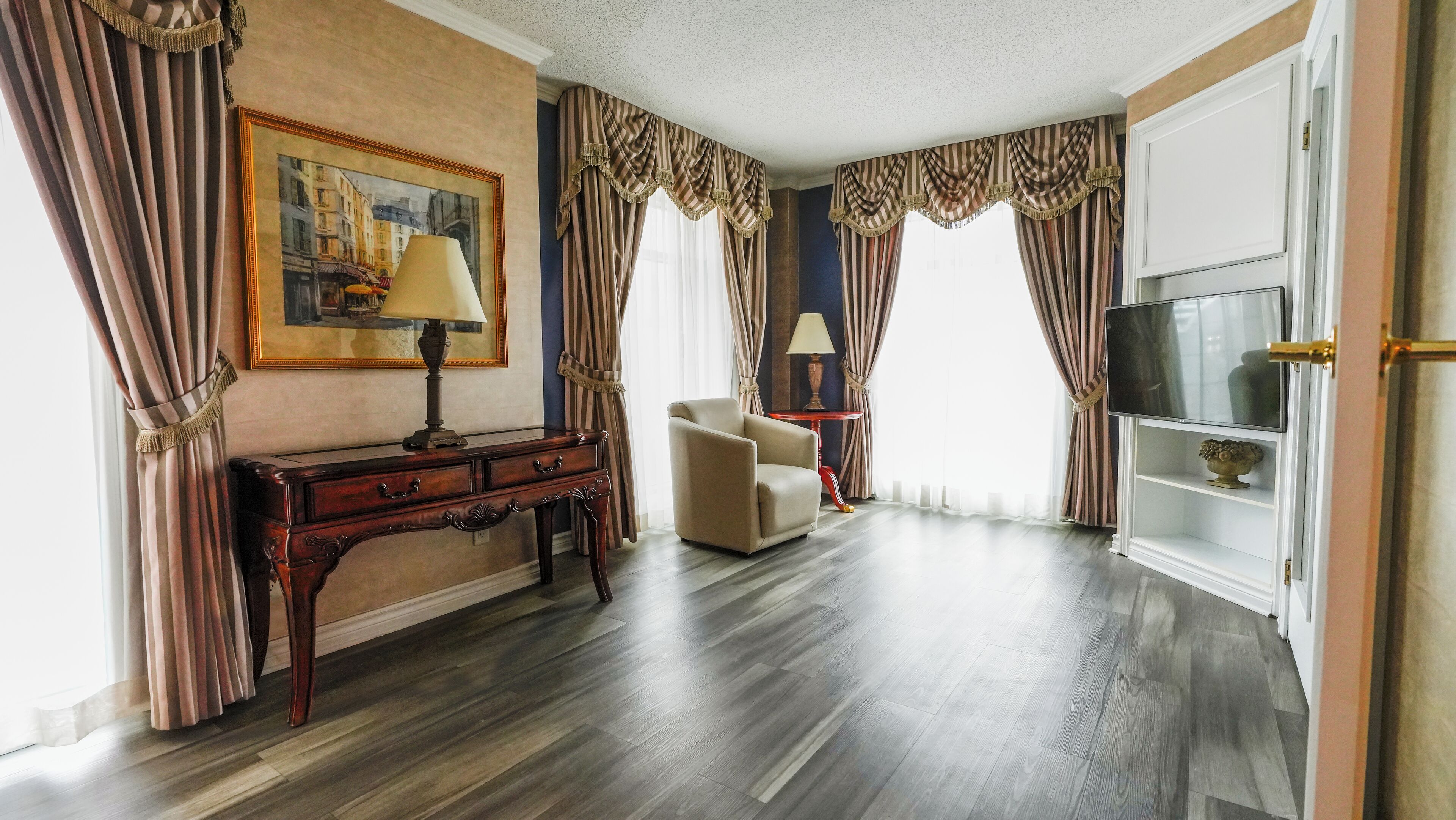 A hotel room with a wooden floor, a desk with a lamp, a chair, a television on a stand, and curtains.