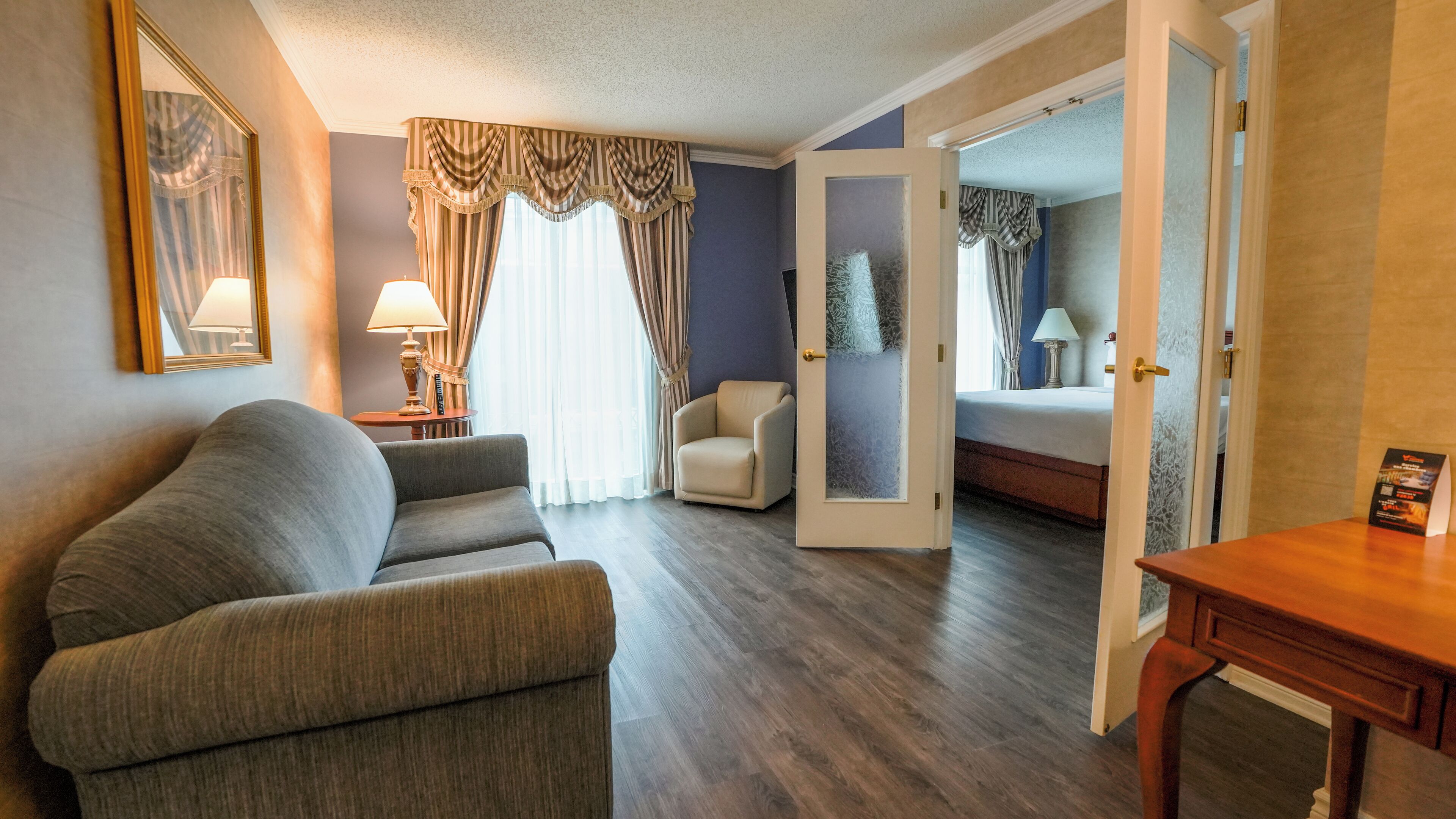 Habitación de hotel con sofá gris, un sillón pequeño, una mesita de madera y una cama con cabecera azul.