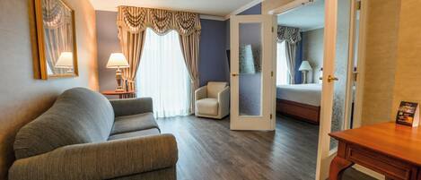 Habitación de hotel con sofá gris, un sillón pequeño, una mesita de madera y una cama con cabecera azul.