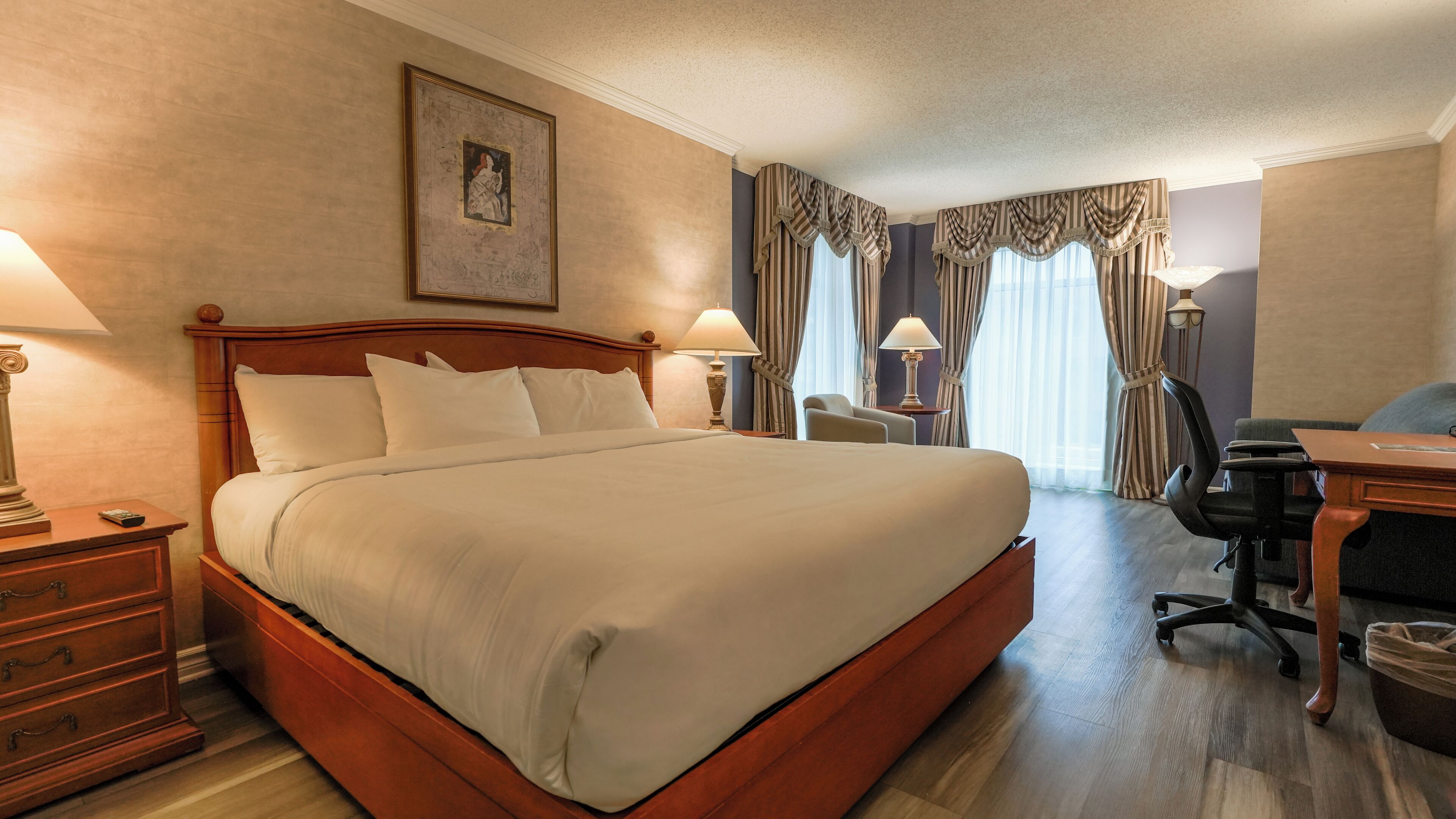 A hotel room with a large bed, two bedside tables with lamps, a desk with a chair, a waste bin, and a framed picture on the wall.