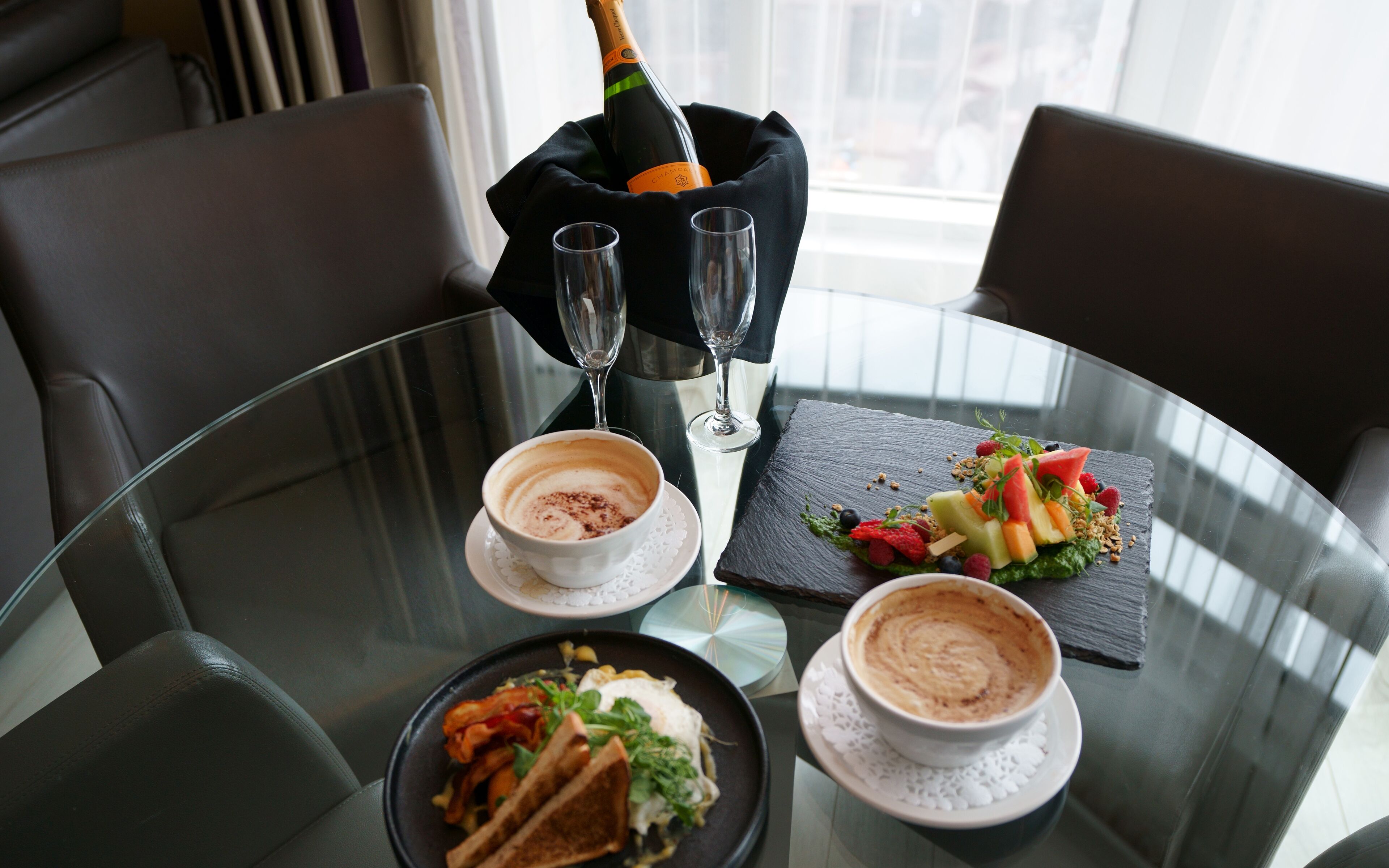 A dining table with a bottle of champagne, two glasses, and two cups of coffee. There is a plate of food and a dessert on the table.