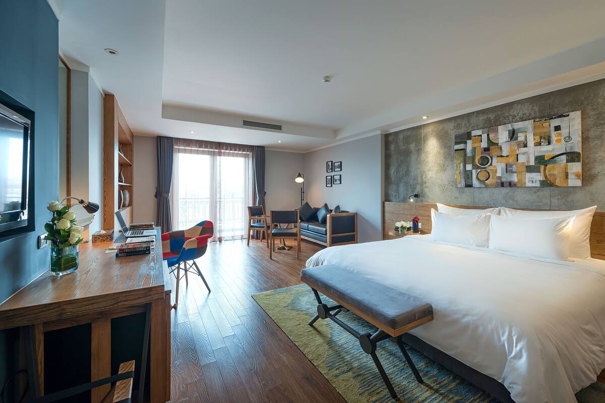 A modern hotel room with a large bed, a desk with a laptop, a chair, a sofa, and a large abstract painting on the wall.