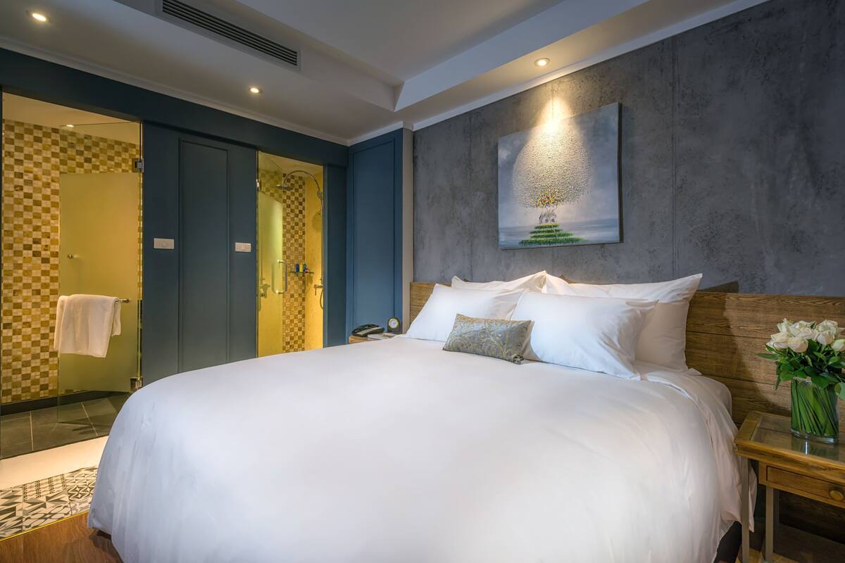 A hotel room with a large bed, a wooden headboard, a nightstand with a vase of flowers, a painting on the wall, and a bathroom with a checkered shower area.