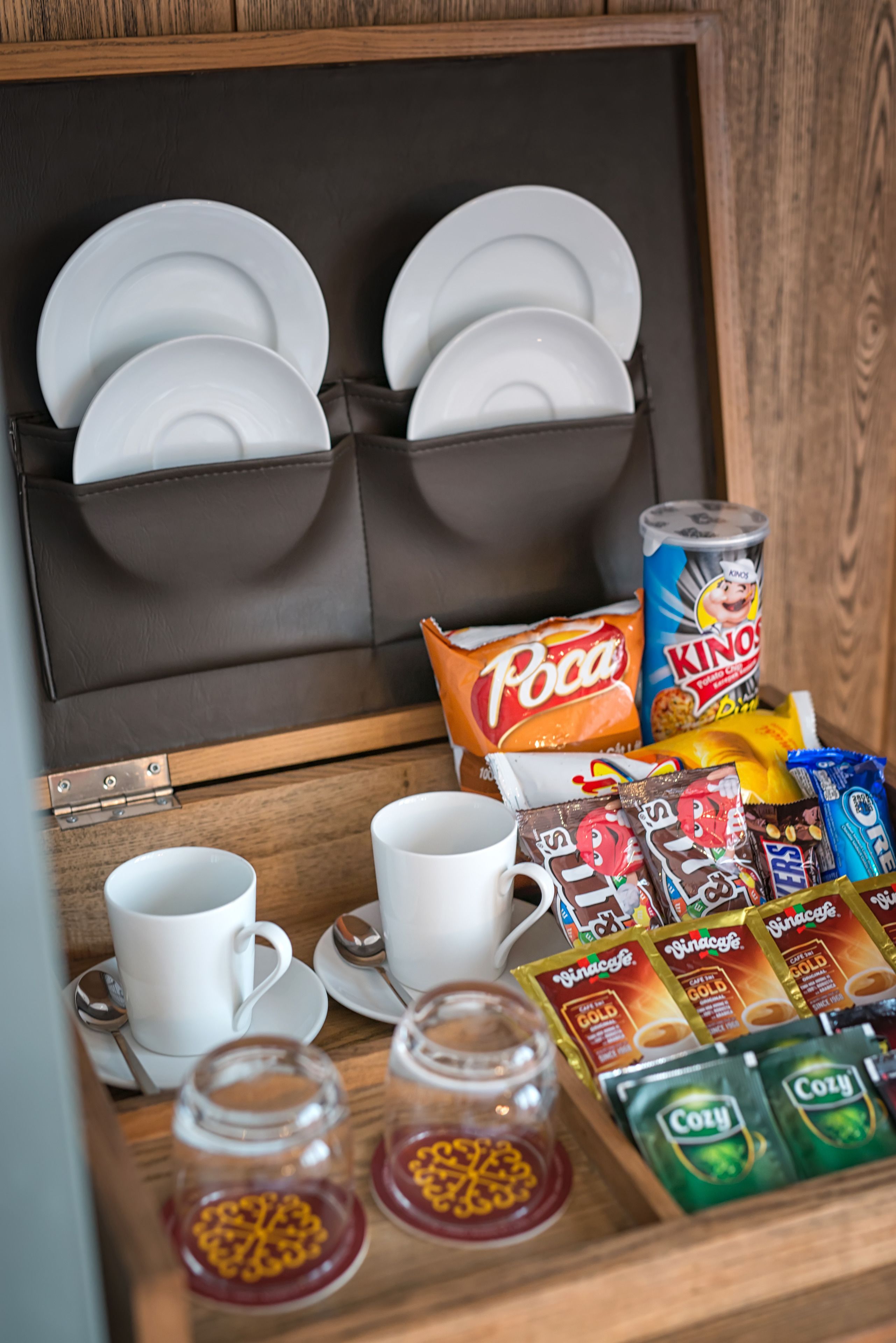 A wooden cabinet with dishes, cups, snacks, and a can of soda.