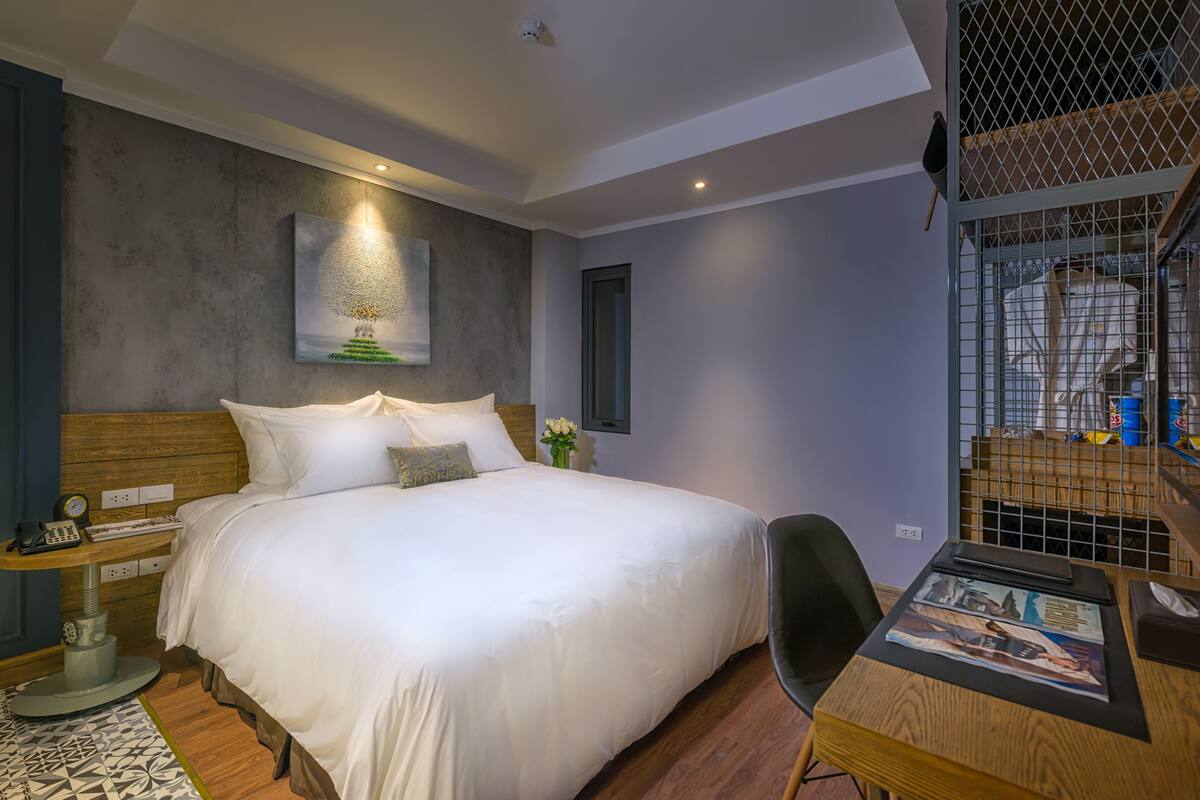 A modern bedroom with a large bed, a bedside table with a lamp, a small desk with a chair, and a wall-mounted TV.