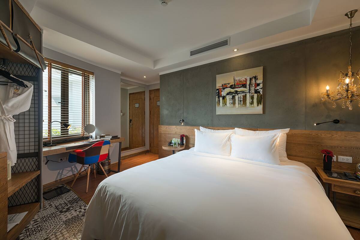 A hotel room with a bed, desk, chair, and a painting on the wall.