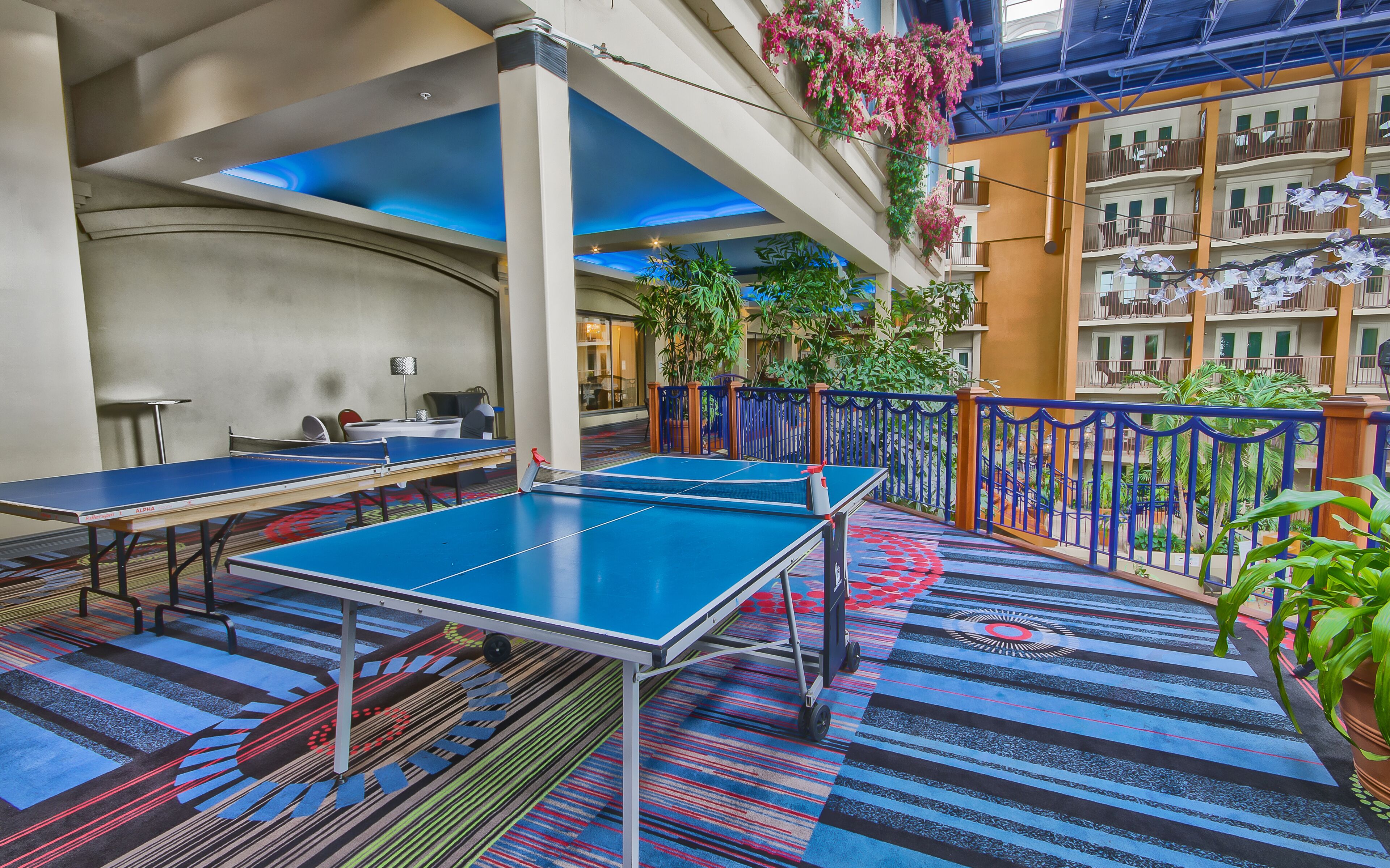 A ping pong table in a hotel lobby with a colorful carpet, a balcony with plants, and a view of the building's interior.