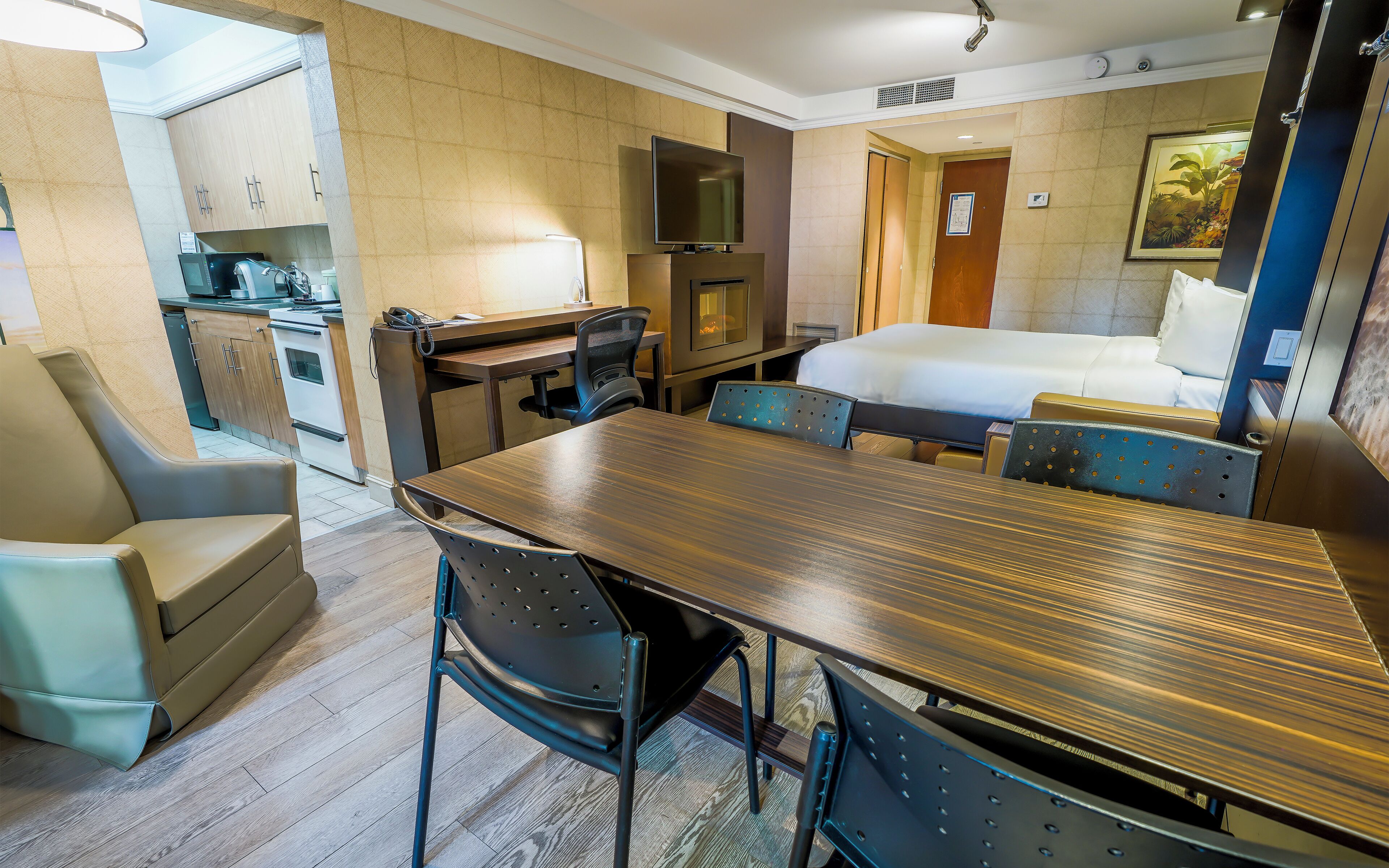 A hotel room with a wooden dining table, two chairs, a bed, a TV, and a kitchenette.