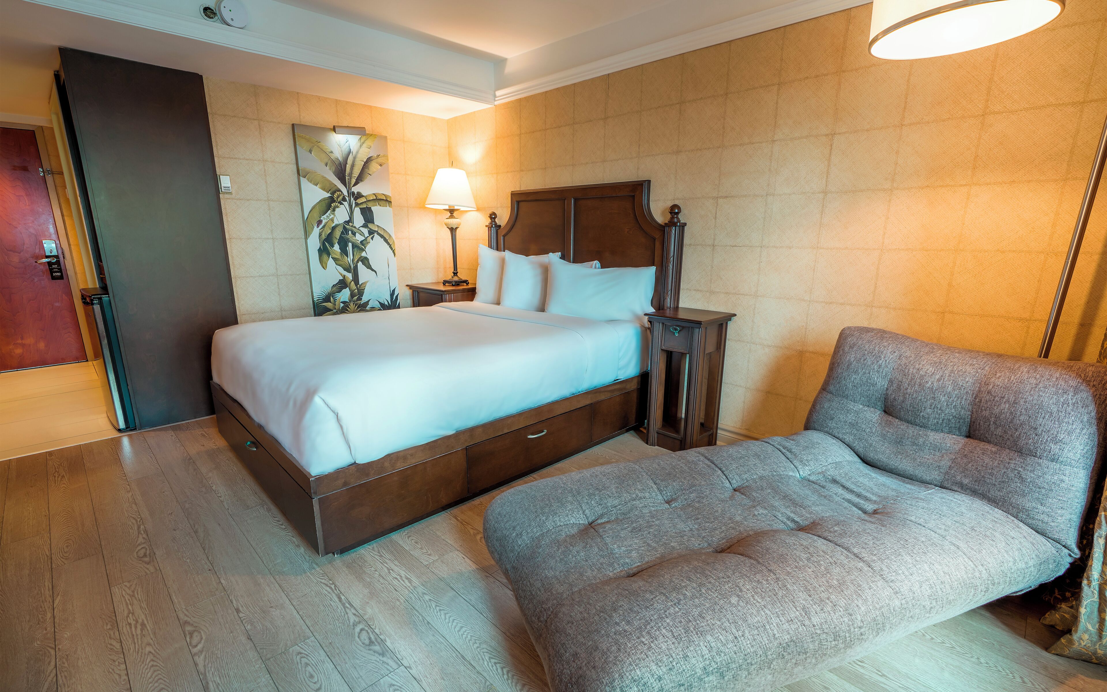 A hotel room with a large bed, a grey armchair, a bedside table with a lamp, and a wall-mounted artwork.