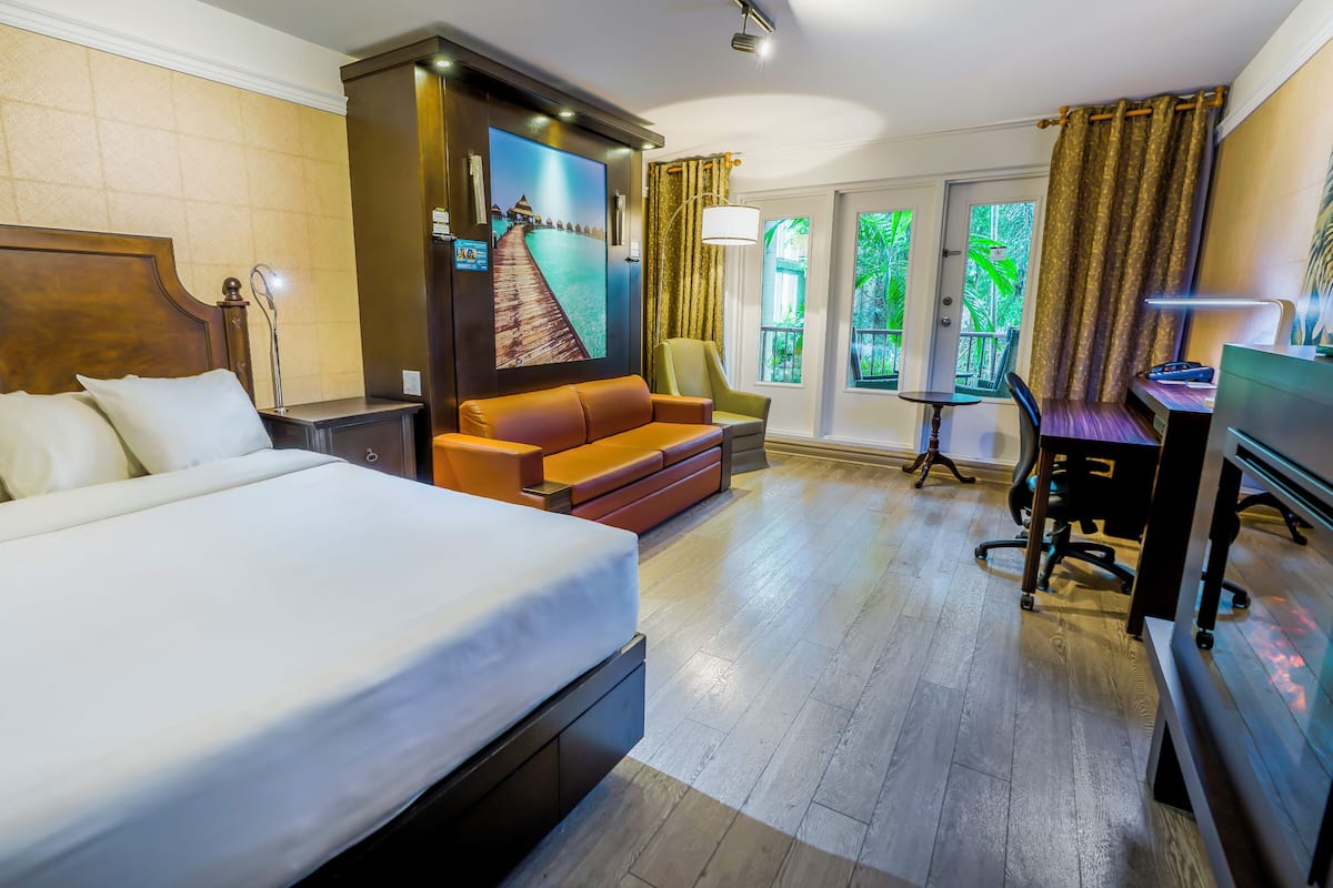 A hotel room with a large bed, a sofa, a desk, and a chair. There are windows with curtains, a painting on the wall, and a lamp.