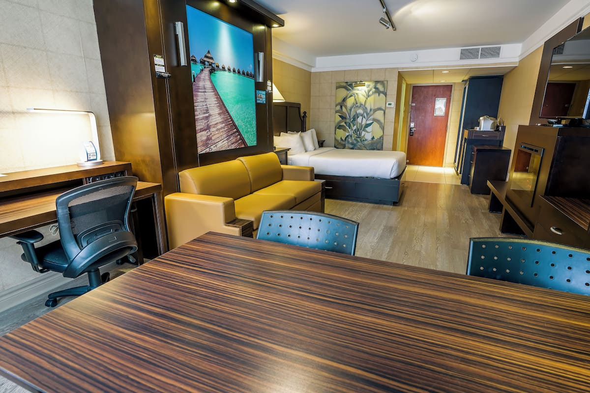 A hotel room with a wooden dining table, a sofa, a bed, a desk with a chair, a television, and artwork on the walls.