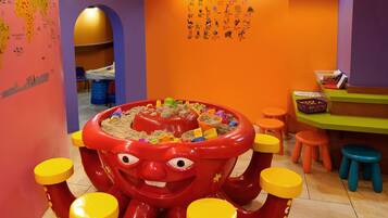 Un espace de jeux pour enfants avec une grande structure de jeu en forme de poulpe rouge, des tabourets colorés et des affiches éducatives accrochées au mur.