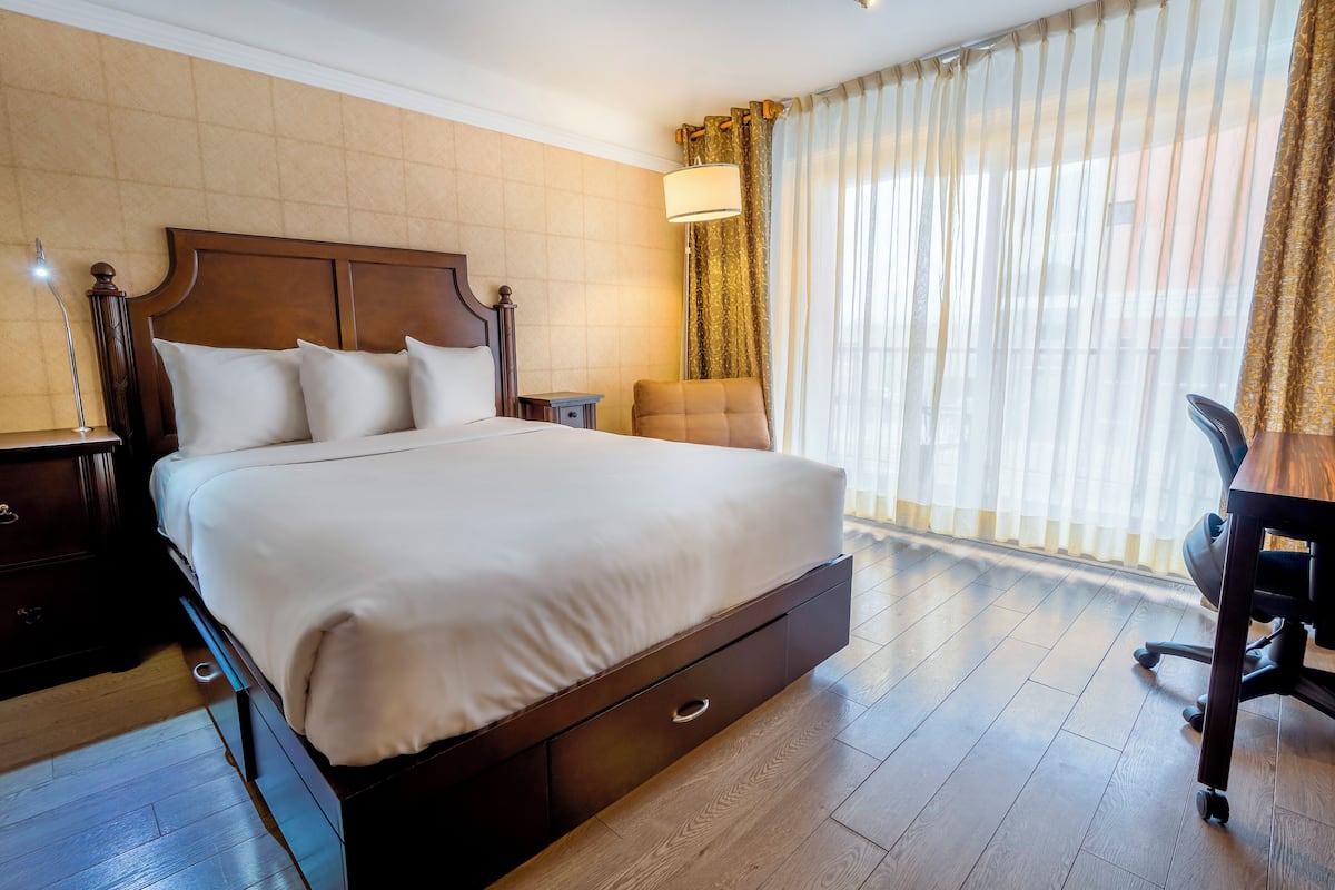 A hotel room with a large bed, wooden headboard, a desk with a chair, a nightstand, and a lamp.