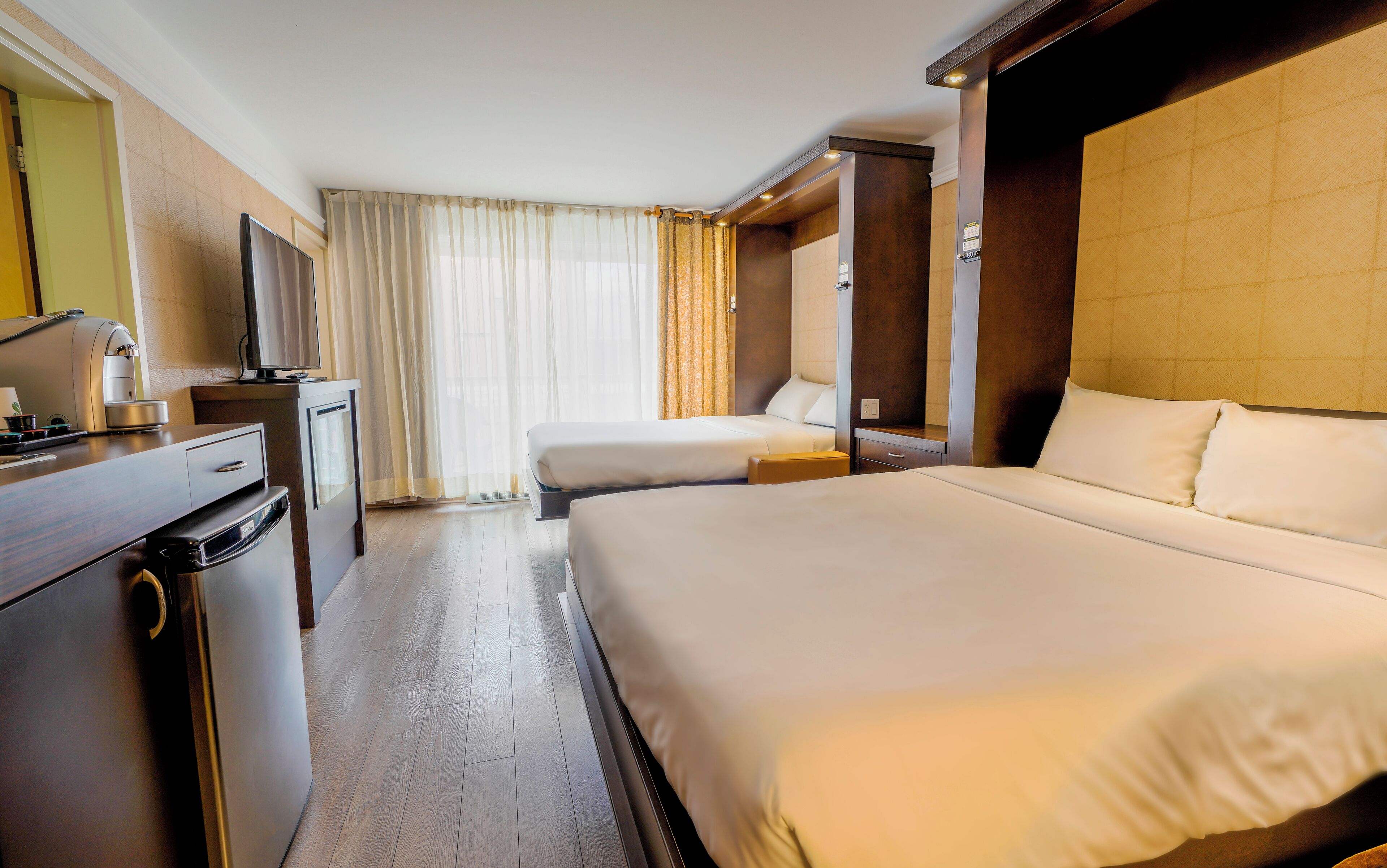 A hotel room with a large bed, a smaller bed, a desk with a coffee machine, and a television.