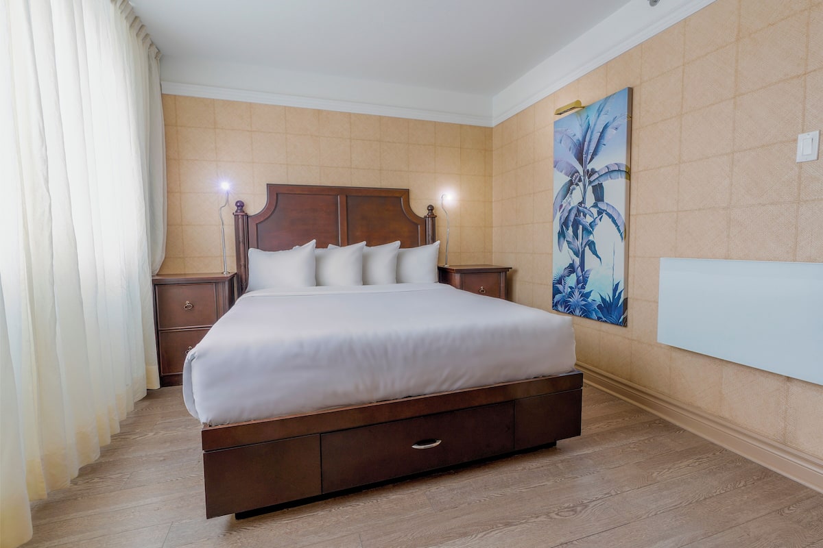 A neatly made bed in a hotel room with a wooden headboard, a nightstand with a lamp, and a large painting on the wall.