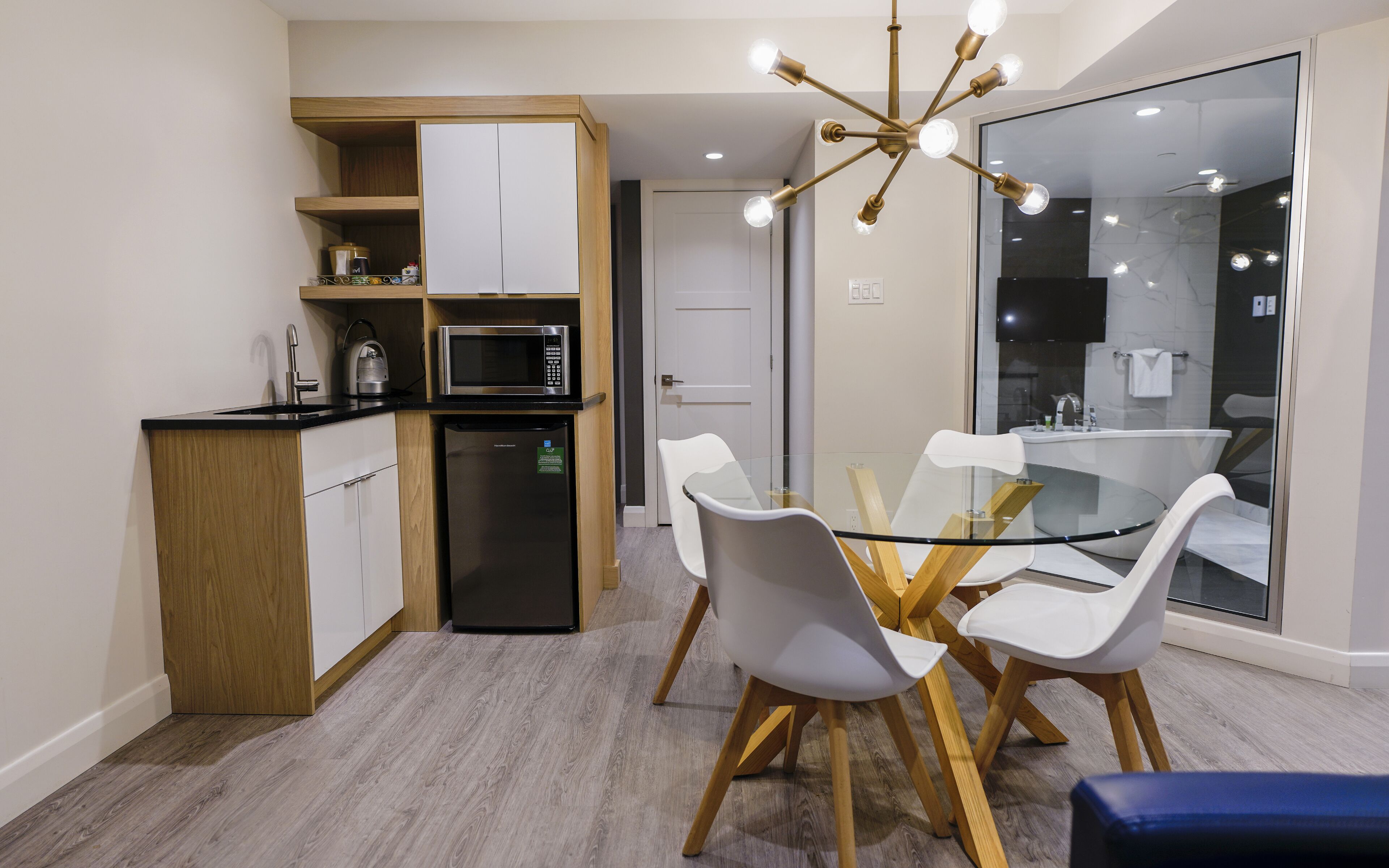 A modern kitchen with a built-in microwave, a refrigerator, and a wooden cabinet. There is a glass dining table with white chairs and a blue bench. A chandelier hangs above the table.