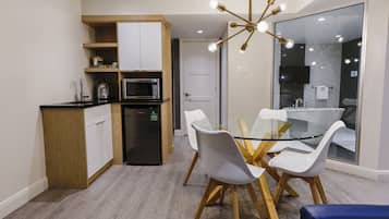 A modern kitchen with a built-in microwave, a refrigerator, and a wooden cabinet. There is a glass dining table with white chairs and a blue bench. A chandelier hangs above the table.