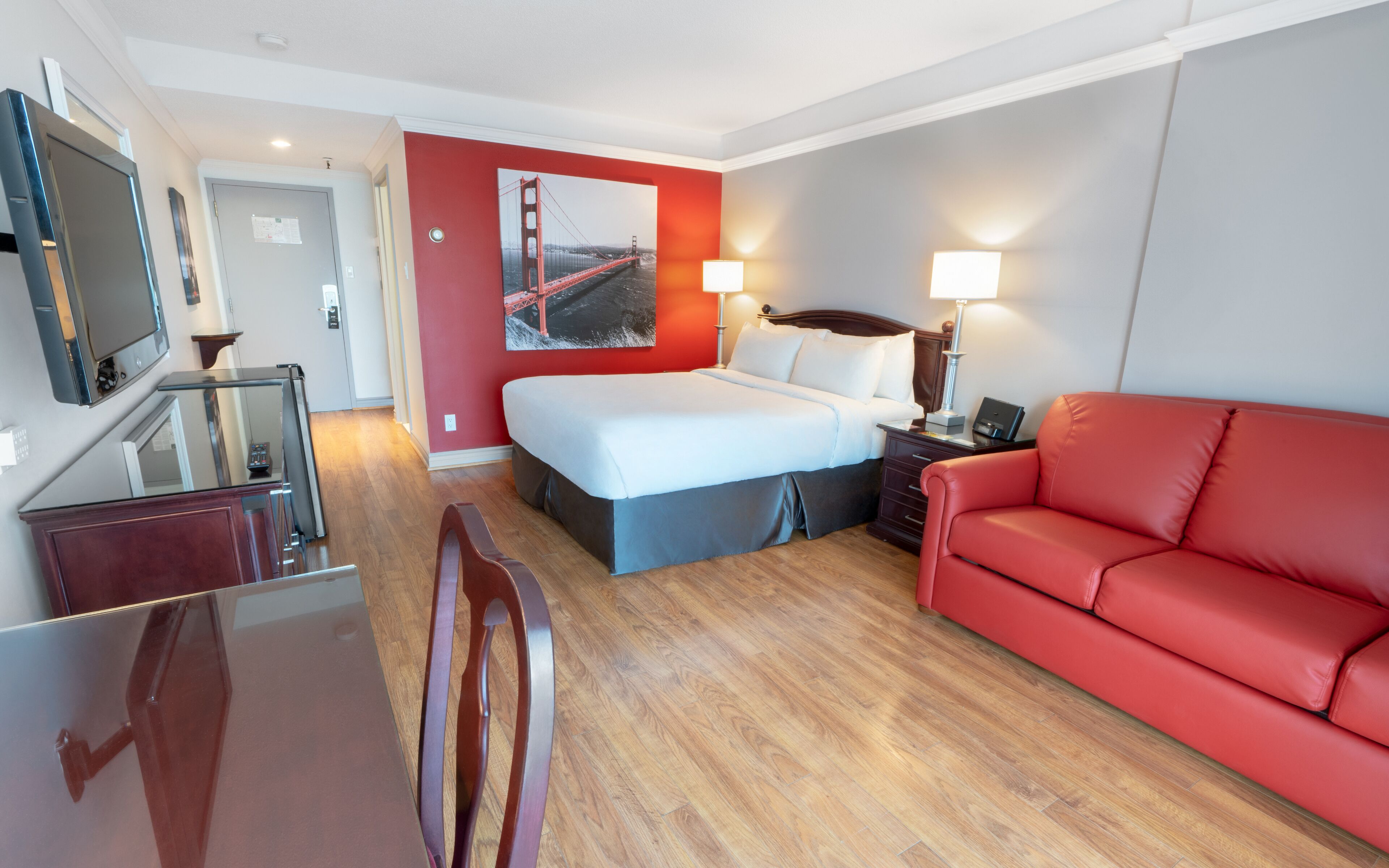 Habitación de hotel con cama, sofá rojo, escritorio con espejo, televisor y un cuadro de un puente.