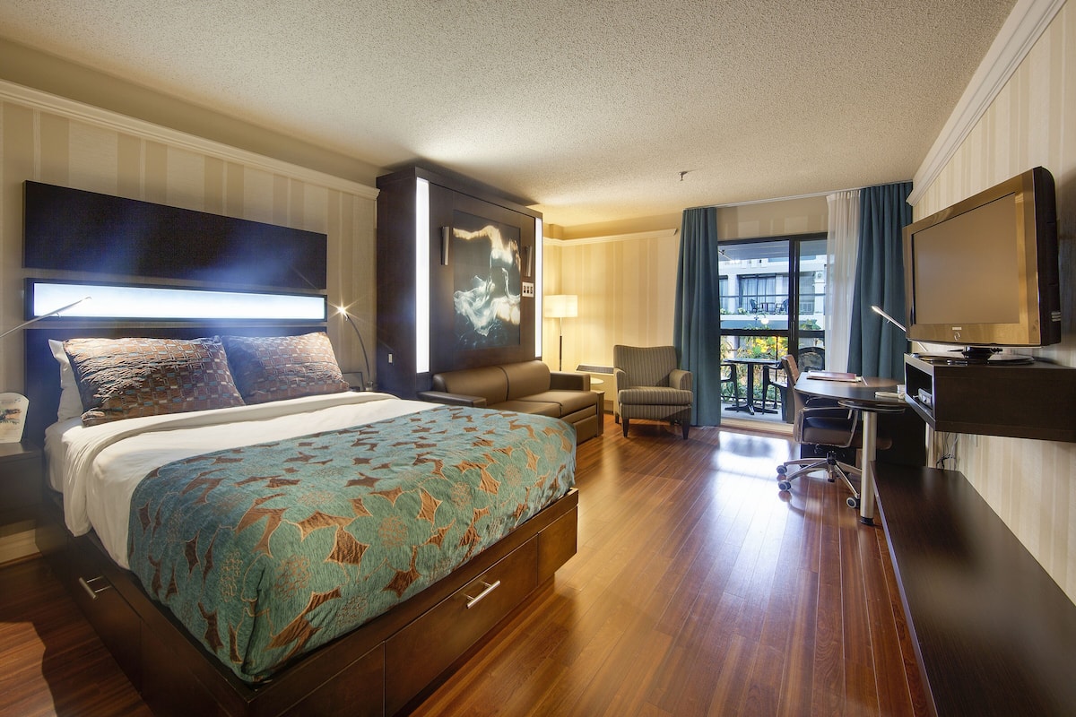 A hotel room with a bed, a desk, a chair, a TV, and a balcony with a view.