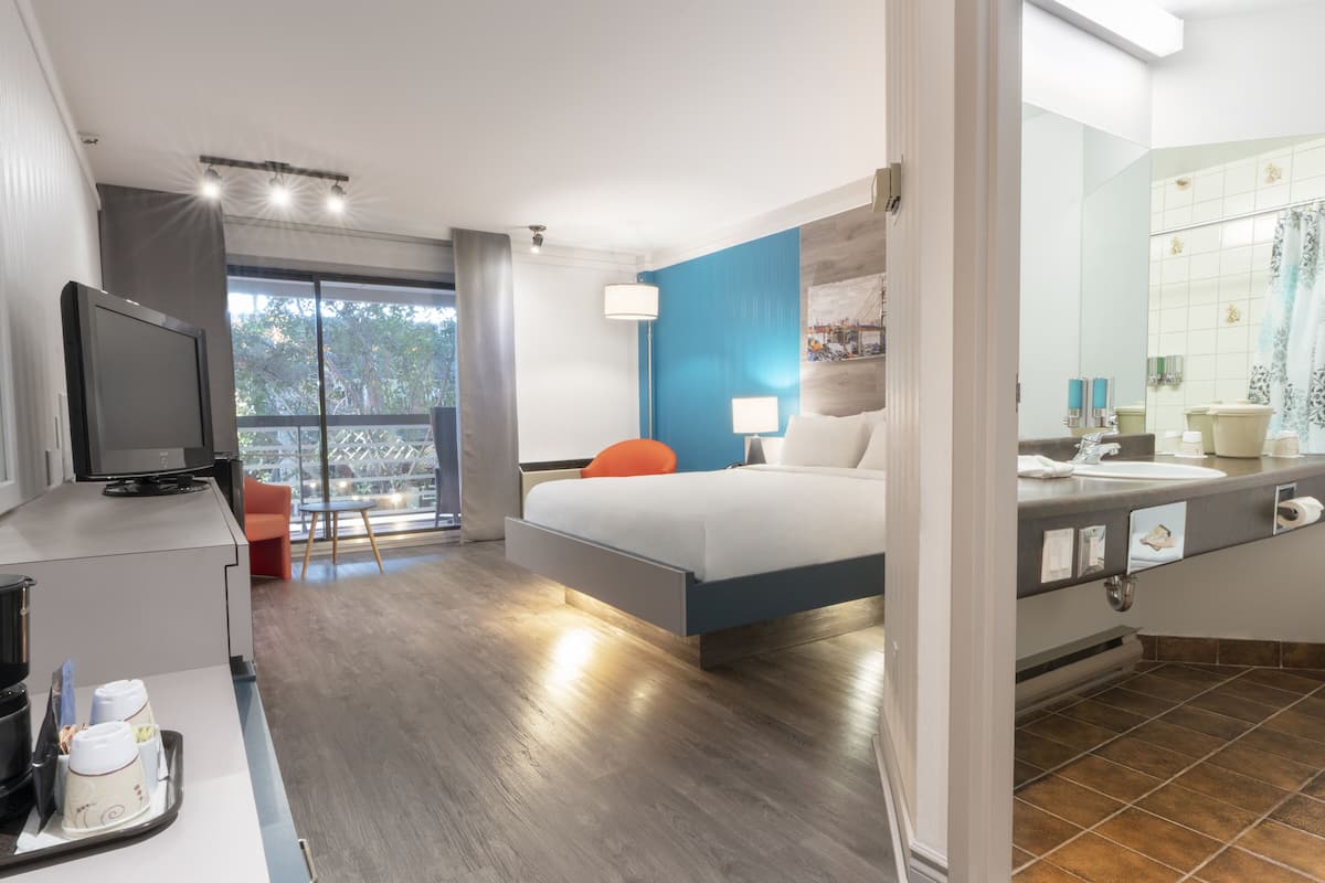 A hotel room with a bed, a desk with a computer, a bathroom with a mirror and sink, and a view of trees through the window.