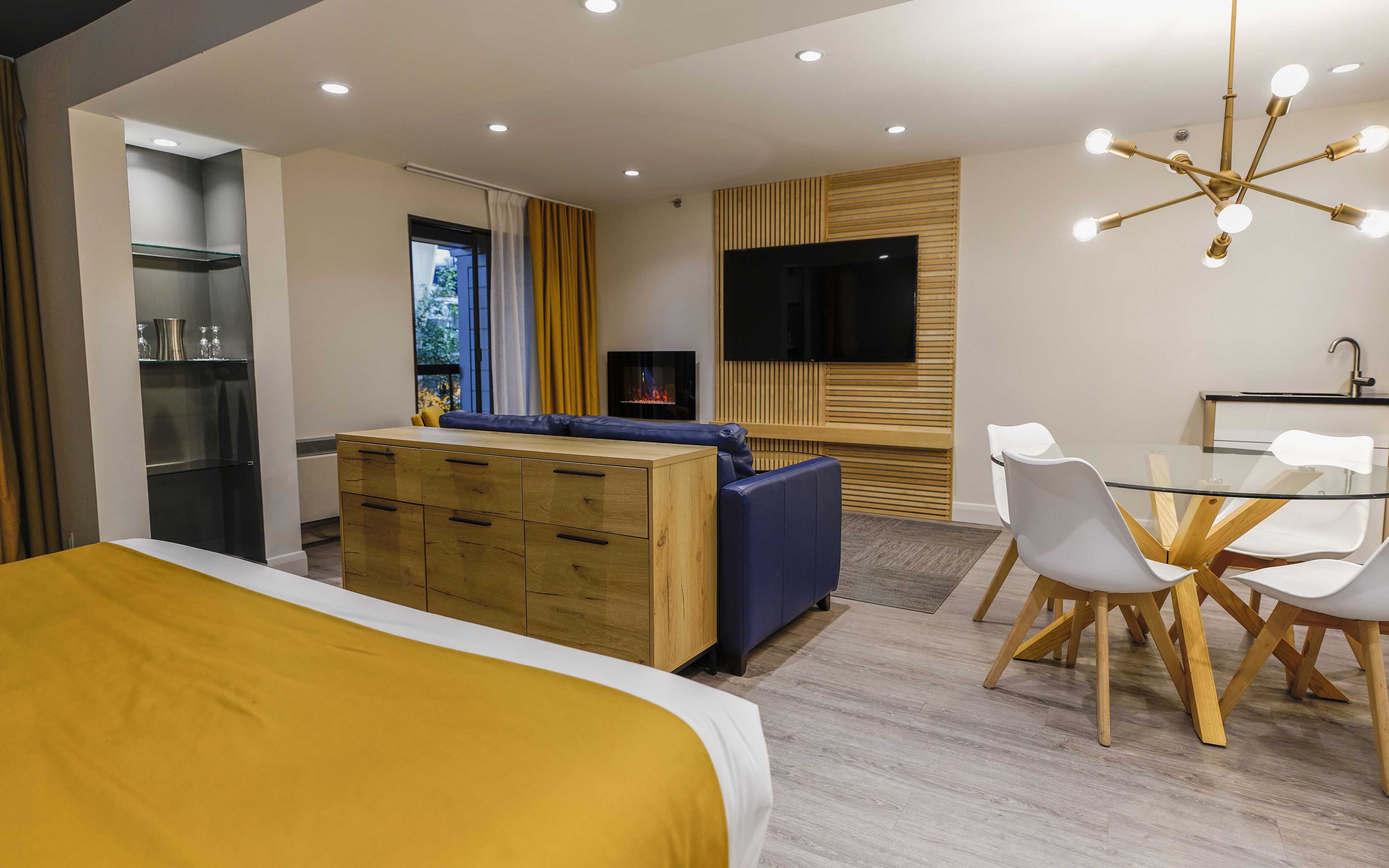 A modern hotel room with a bed, a wooden dresser, a TV, a dining area with a glass table and chairs, and a ceiling light fixture.