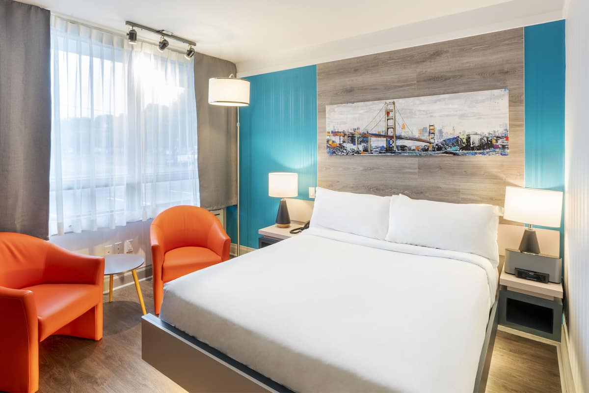 A hotel room with a large bed, two orange chairs, a small table, a painting of a bridge, and a nightstand with a lamp.