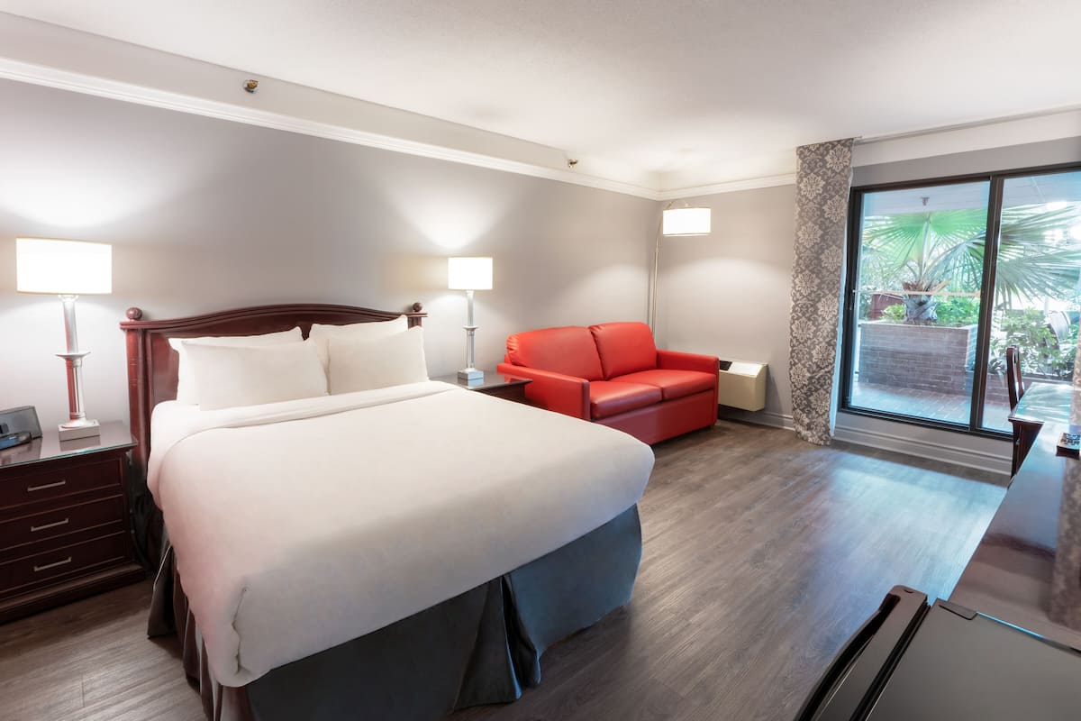 A hotel room with a large bed, a red sofa, a desk with a lamp, and a view of the outdoors through a large window.