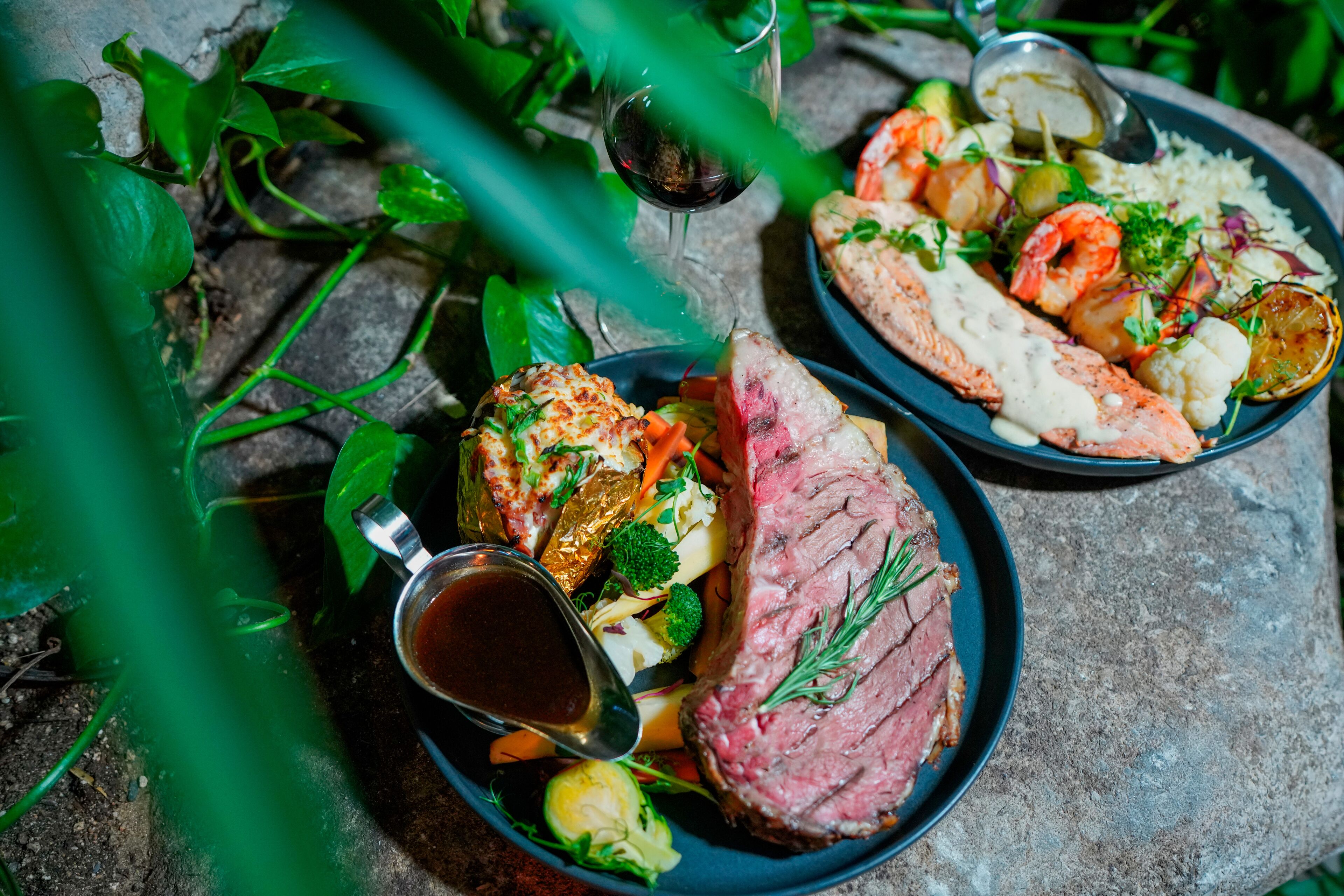 Un plat composé de steak, de crevettes et de légumes, accompagné d’un verre de vin rouge et d’un petit ramequin de sauce.