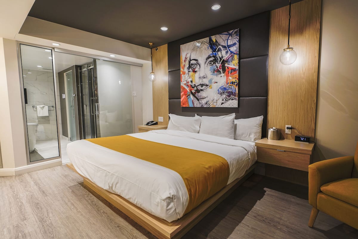 A modern hotel room with a large bed, a glass shower enclosure, a wooden headboard, and a digital clock on the nightstand.