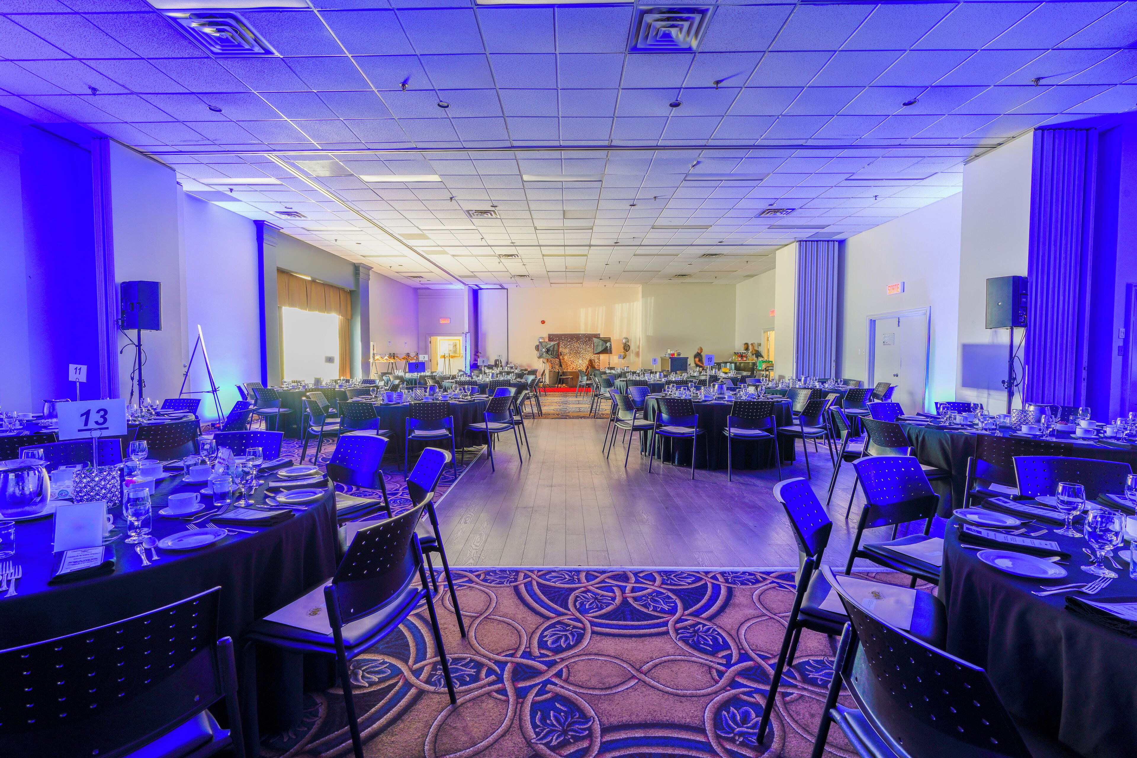 A banquet hall with multiple round tables, chairs, and a stage at the far end.