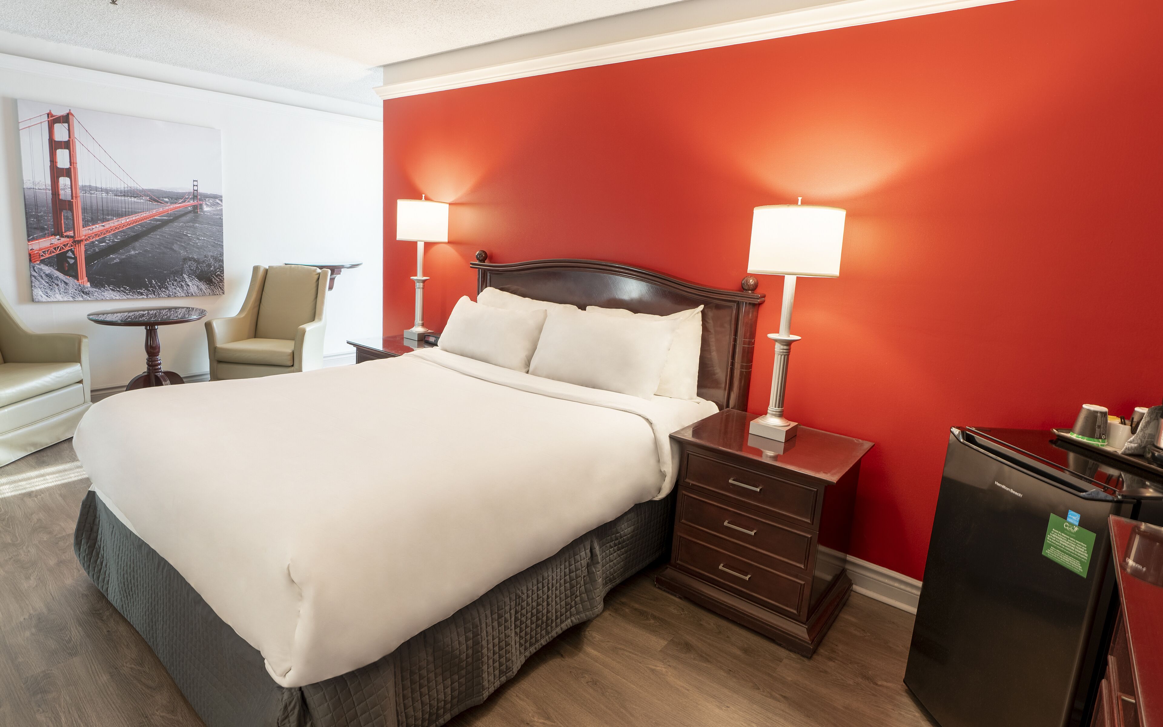 Habitación de hotel con una cama grande, dos lámparas de noche, una mesita de noche, un escritorio, una silla y un refrigerador.
